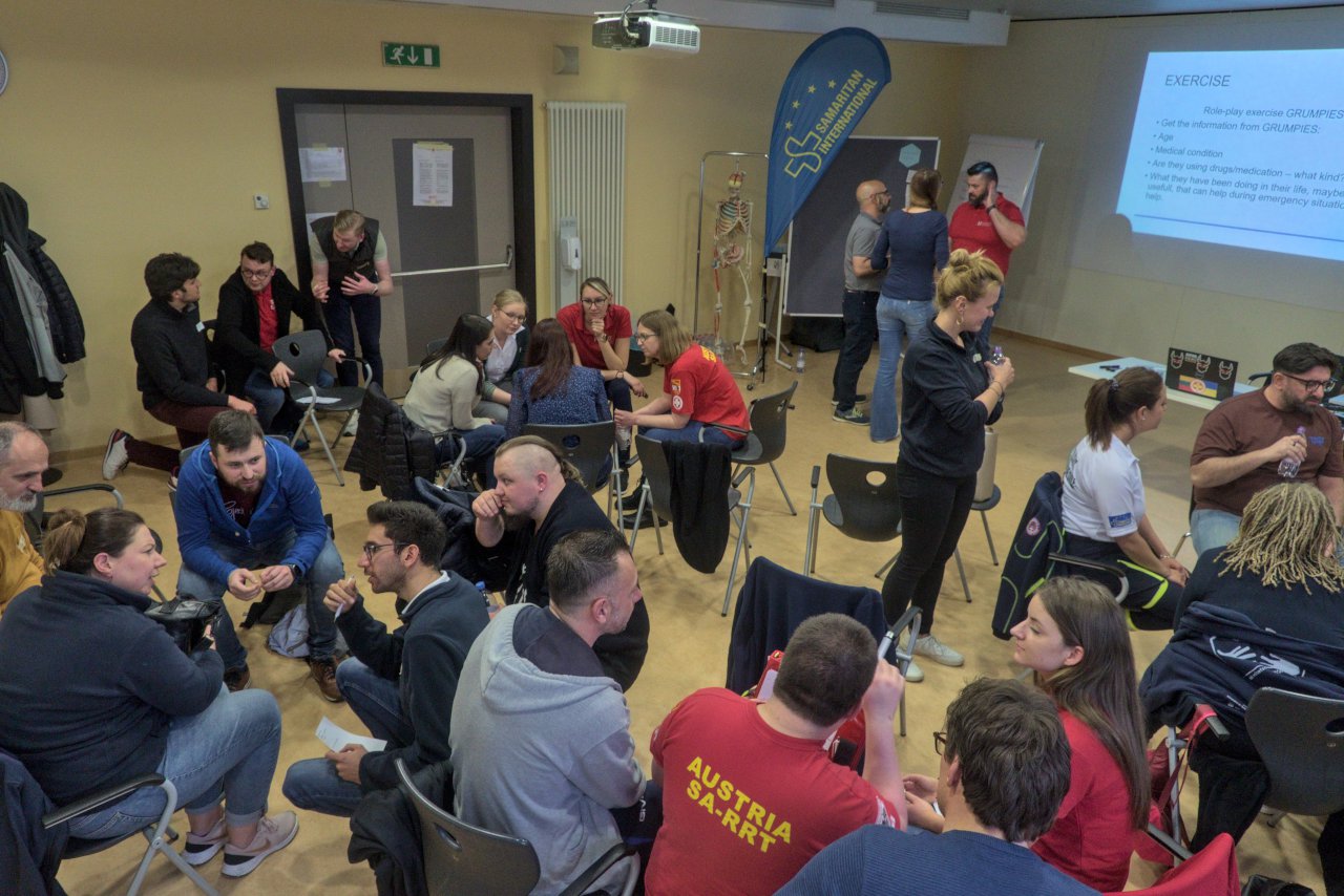 Classroom scene during the I_TEM workshop. A group of some 30 people is sitting or standing divided into smaller group circles and discussing a group work exercise.