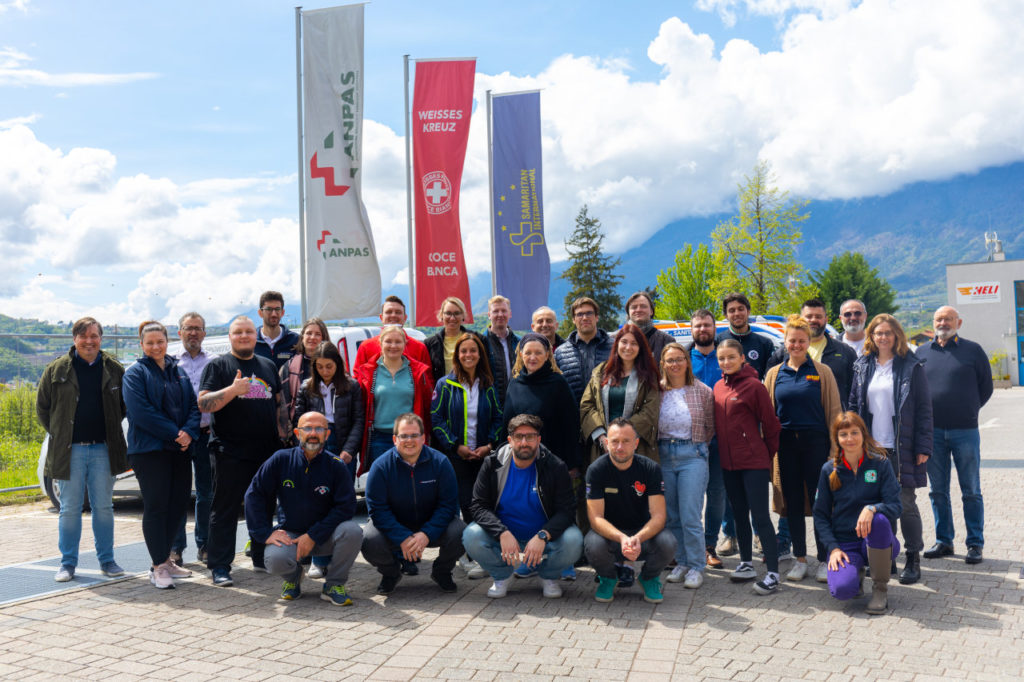Group photo of the participant and trainers at the I_TEM C2 meeting. The group is in a parking lot. Behind them, the three flags (from left to right) of ANPAS, White Cross and SAM.I. can be seen. The background is a mountainous vista. The sky is sunny but with some large white clouds in the distance.