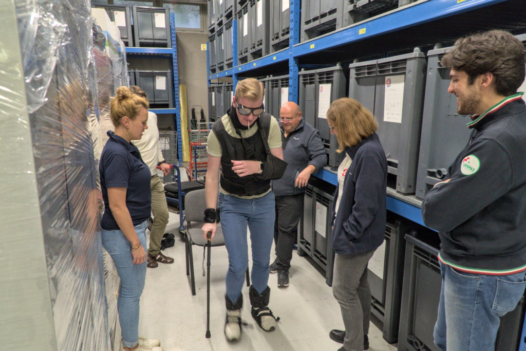 A practical exercise at the I_TEM C2 training. One participant is wearing an aging simulation suit and is walking using a cane while five other people look on. The scene is set in a warehouse corridor, the person in the aging suit is walking from a chair from which he started towards the camera.